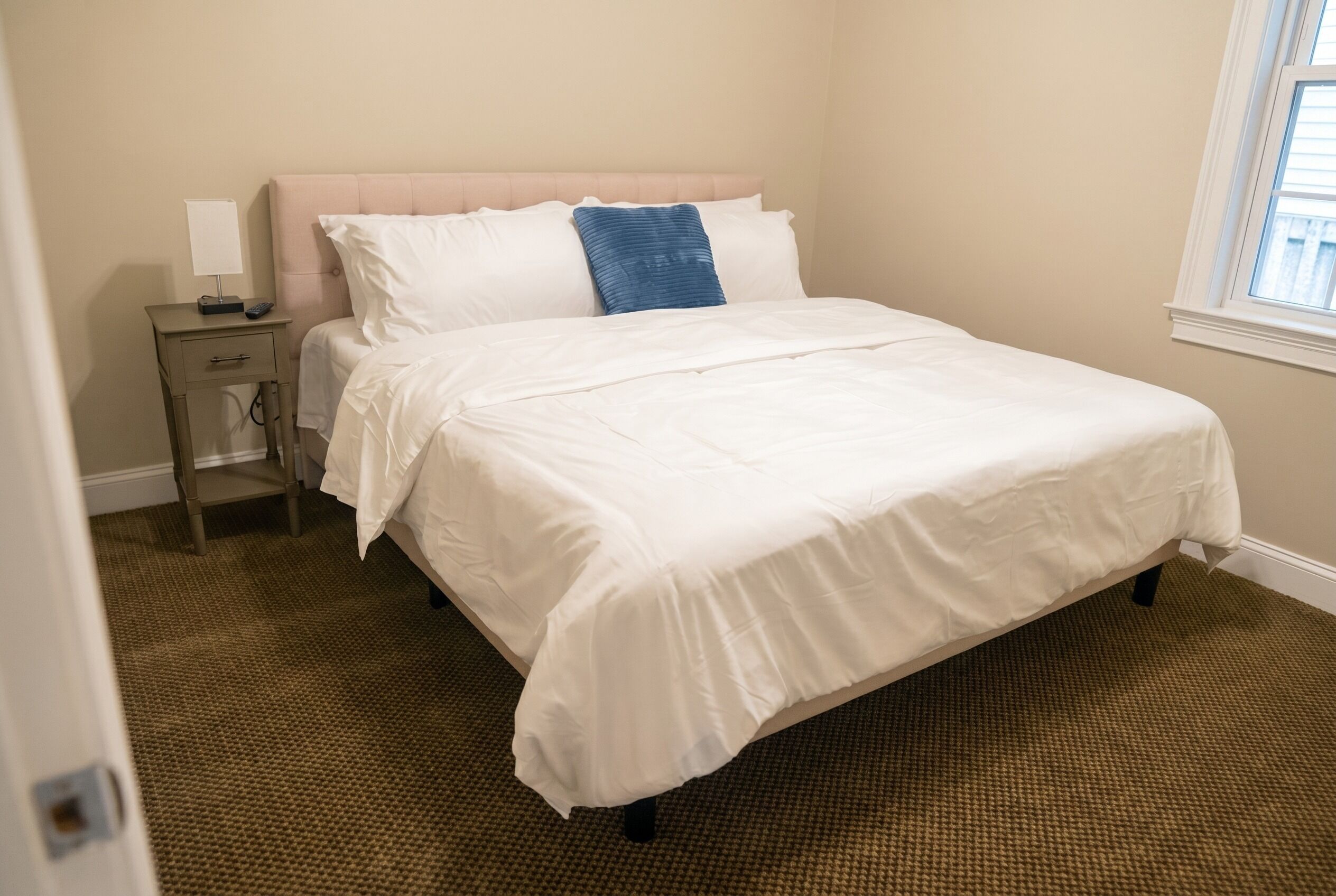 A neatly made bed with a headboard, a bedside table with a lamp, and a window with blinds.