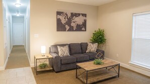 Uma sala de estar com um sofá cinzento, uma mesa de centro de madeira, um mapa-múndi na parede, uma planta e uma janela com persianas.