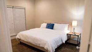 Quarto de hotel com cama, mesa de cabeceira, luminária e janela com persianas.