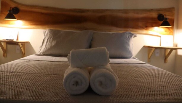 Una cama bien hecha con una toalla enrollada en el centro, dos almohadas y dos lámparas de noche sobre mesitas de madera.