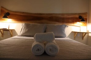Una cama bien hecha con una toalla enrollada en el centro, dos almohadas y dos lámparas de noche sobre mesitas de madera.