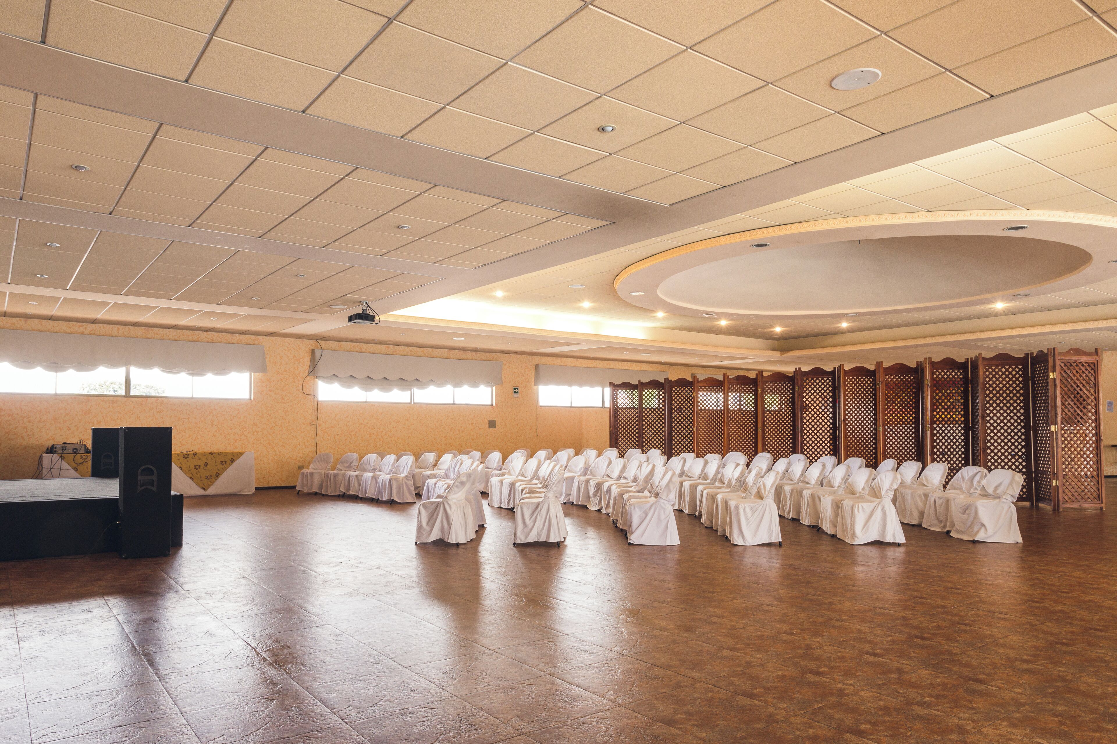Un salón amplio con filas de sillas cubiertas con manteles blancos, un escenario con altavoces y un panel de separación de madera.