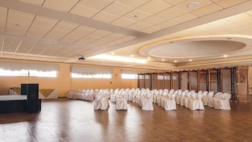 Un salón amplio con filas de sillas cubiertas con manteles blancos, un escenario con altavoces y un panel de separación de madera.