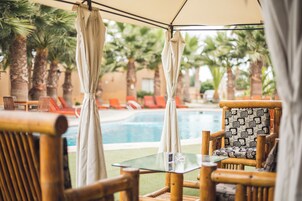 Zona de piscina con sillas de madera y una mesa de vidrio, rodeada de palmeras y una alberca.