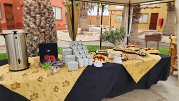 Una mesa de buffet con distintos alimentos y una estación de café, montada al aire libre bajo una carpa.