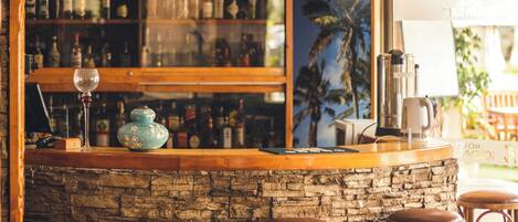Zona de bar con pared de piedra, mostrador de madera y taburetes. Botellas y vasos en las estanterías, y vistas a palmeras a través de una ventana.