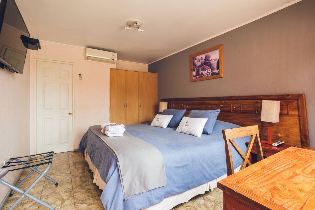 A bedroom with a bed, a wooden wardrobe, a wall-mounted air conditioner, a framed picture, and a wooden nightstand with a lamp.