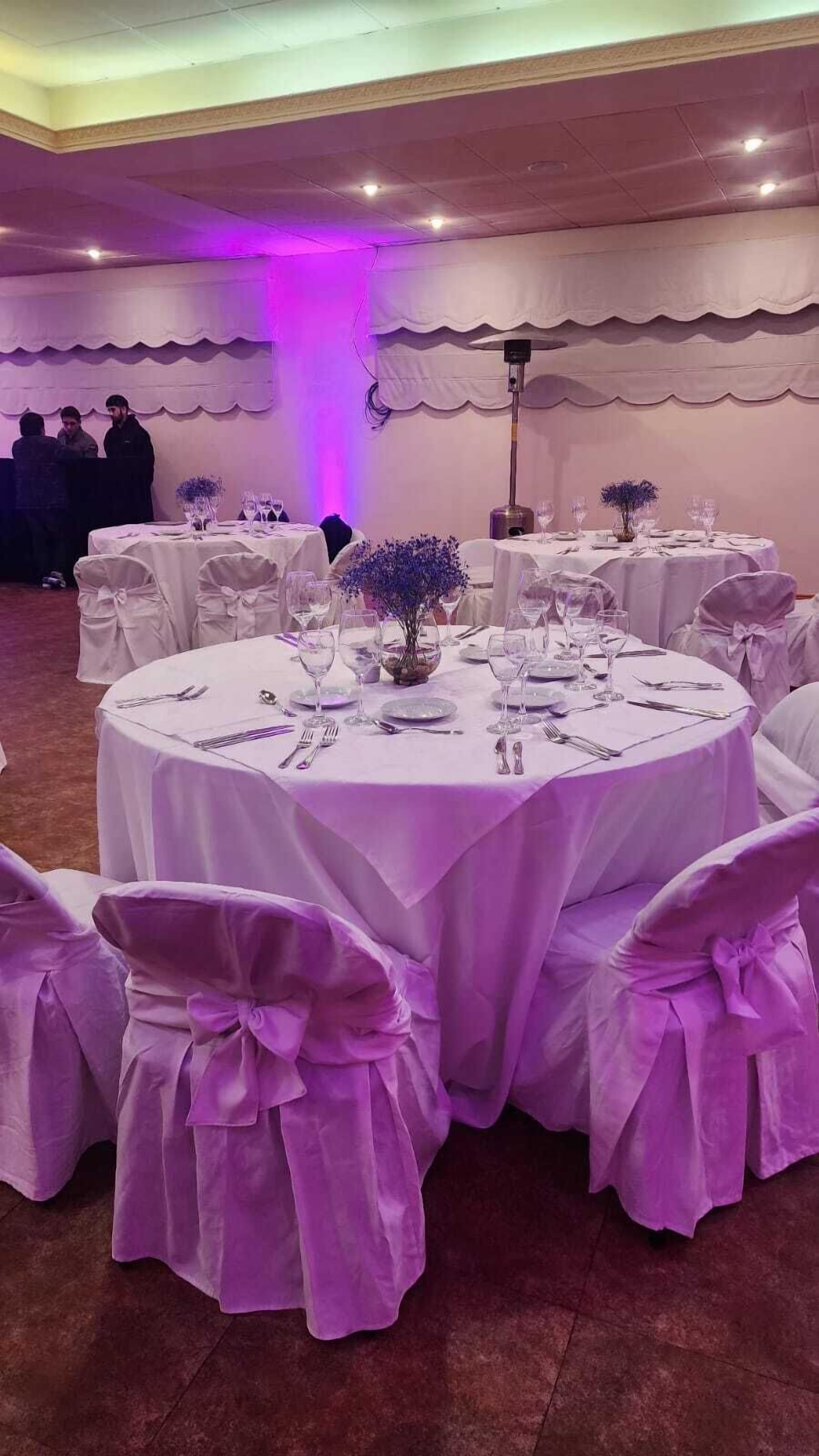 Ein Festsaal mit runden Tischen, gedeckt für eine festliche Veranstaltung, mit weißen Tischdecken und violetten Stühlen.