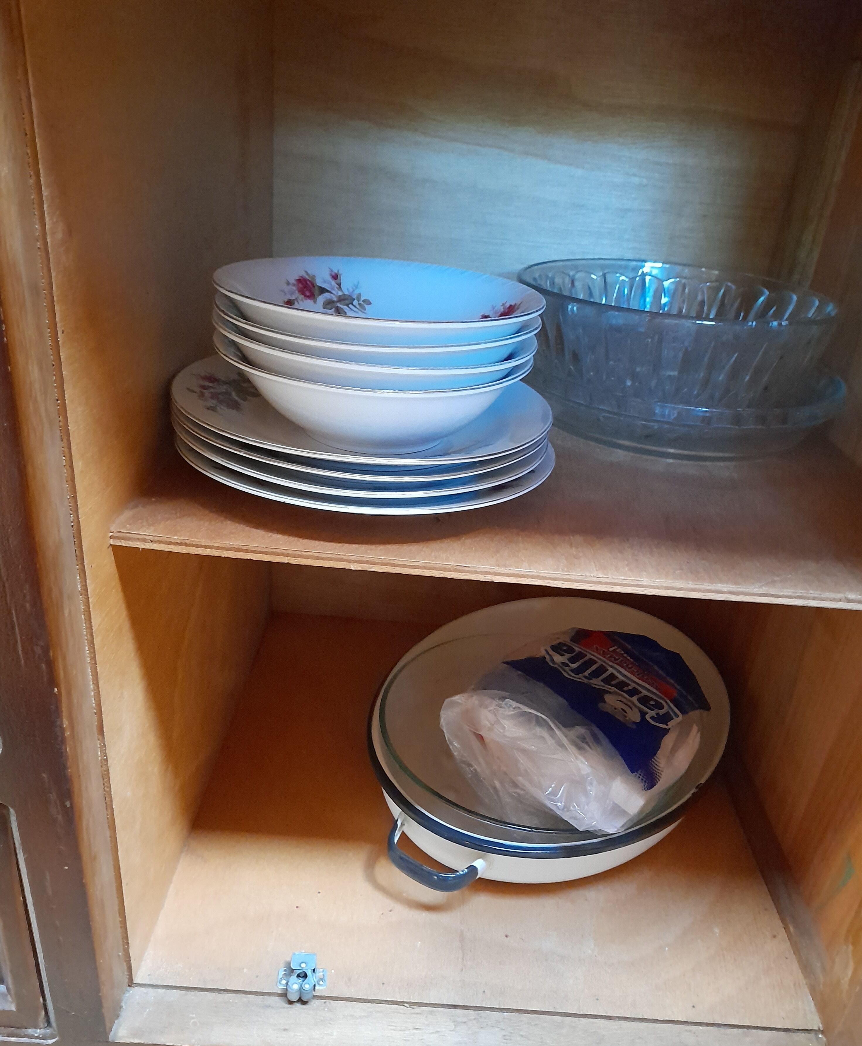 Un meuble en bois doté d’étagères où sont empilées des assiettes, un saladier en verre et un récipient contenant un sac en plastique.