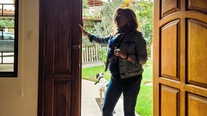 Una puerta de madera con un pestillo metálico, una persona de pie en el umbral y un perro con correa afuera.