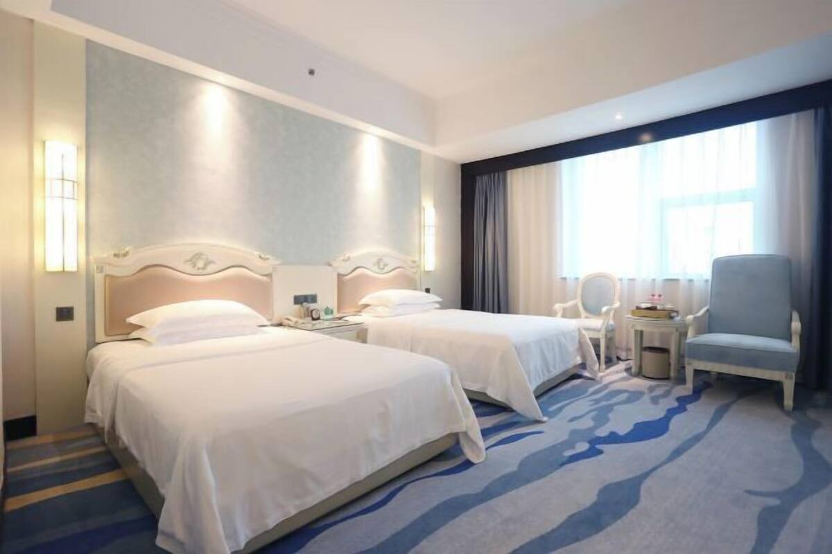 A hotel room with two beds, a small table, a chair, and a large window with curtains.