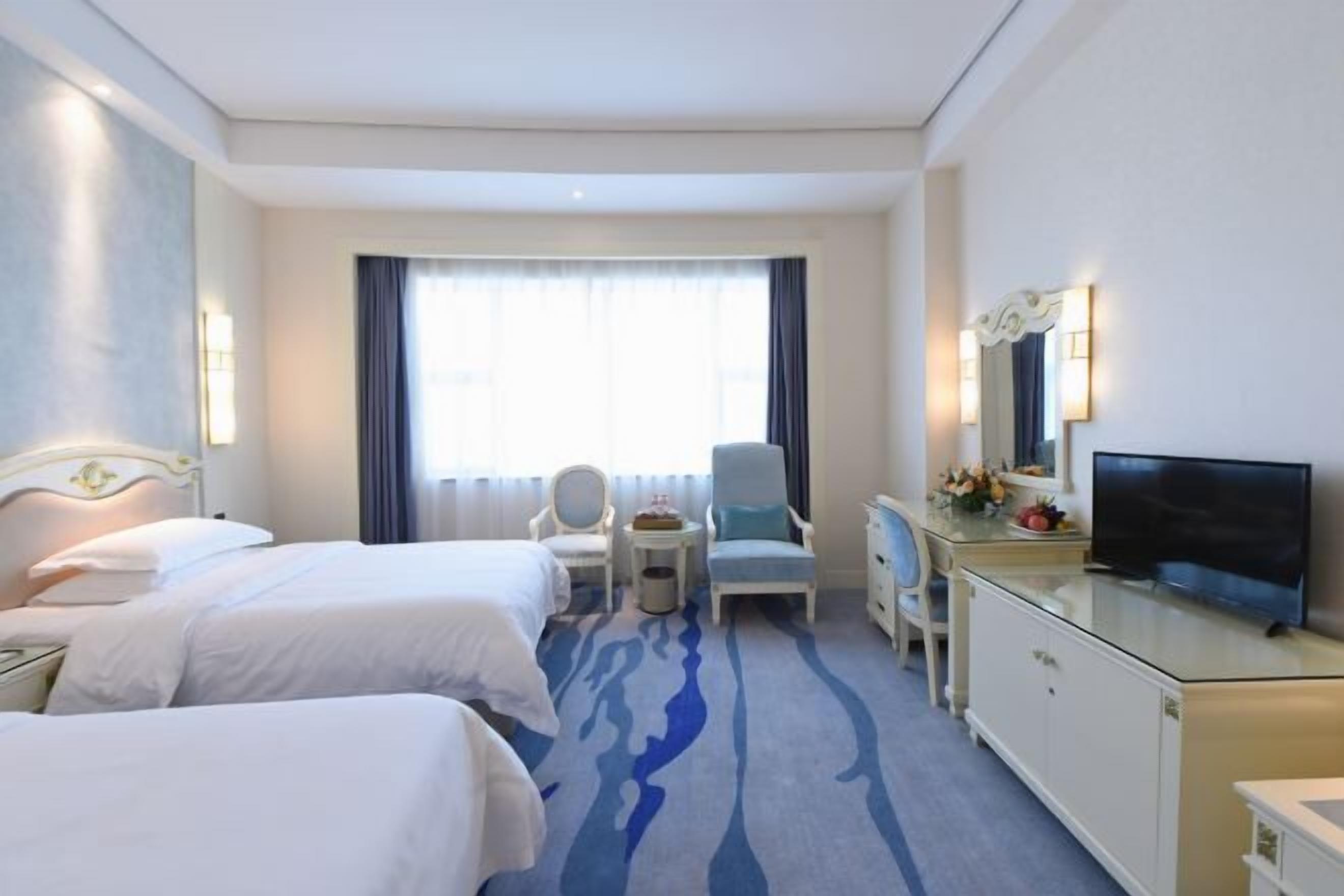 A hotel room with two beds, a desk with a TV, a chair, and a small table with flowers.