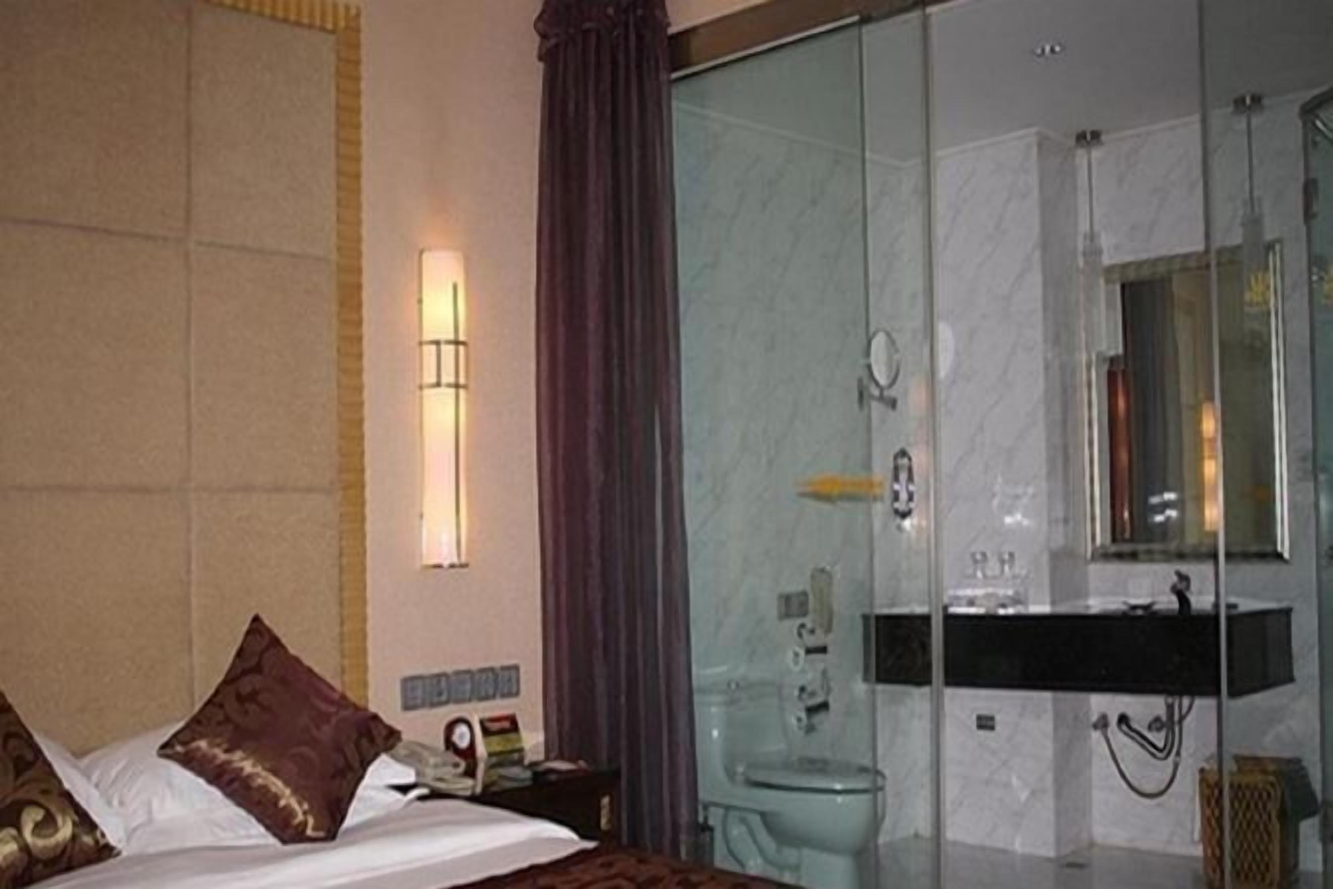 A hotel room with a bed, a bedside table, a mirror, a bathroom with a glass shower enclosure, and a sink.