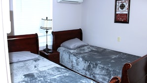 A bedroom with two single beds, a bedside table with a lamp, an air conditioner, and a framed quote on the wall.