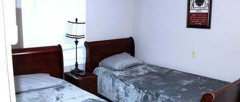 A bedroom with two single beds, a bedside table with a lamp, an air conditioner, and a framed quote on the wall.