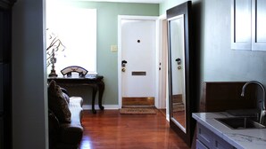 A hallway with a wooden floor, a door, a small table with a lamp, and a kitchen area with a sink.