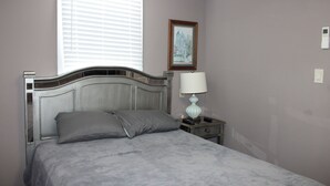 A bedroom with a bed, a bedside table with a lamp, a framed picture on the wall, and a window with blinds.