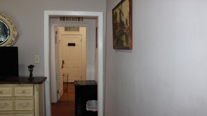 A hallway with a wooden floor, a white door at the end, a small trash bin, and a painting on the wall. - 1 Bedroom Suite upstairs (NEW ORLEANS)