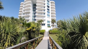 A modern, multi-story hotel with a curved facade, surrounded by lush greenery and a wooden walkway leading to the entrance. - The Pierre 507 by FGC (Longboat Key)