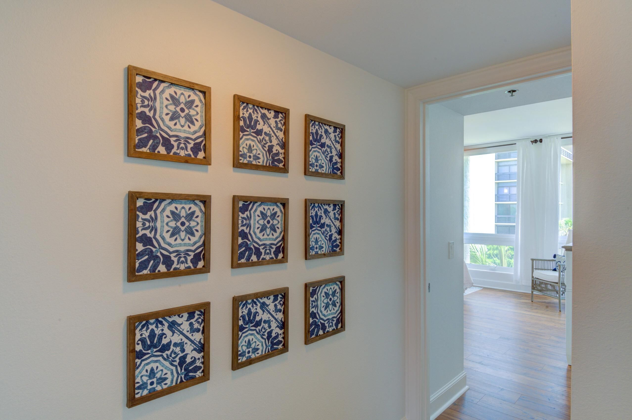 A hallway with nine framed blue and white tiles on the wall, leading to a balcony with a white chair.