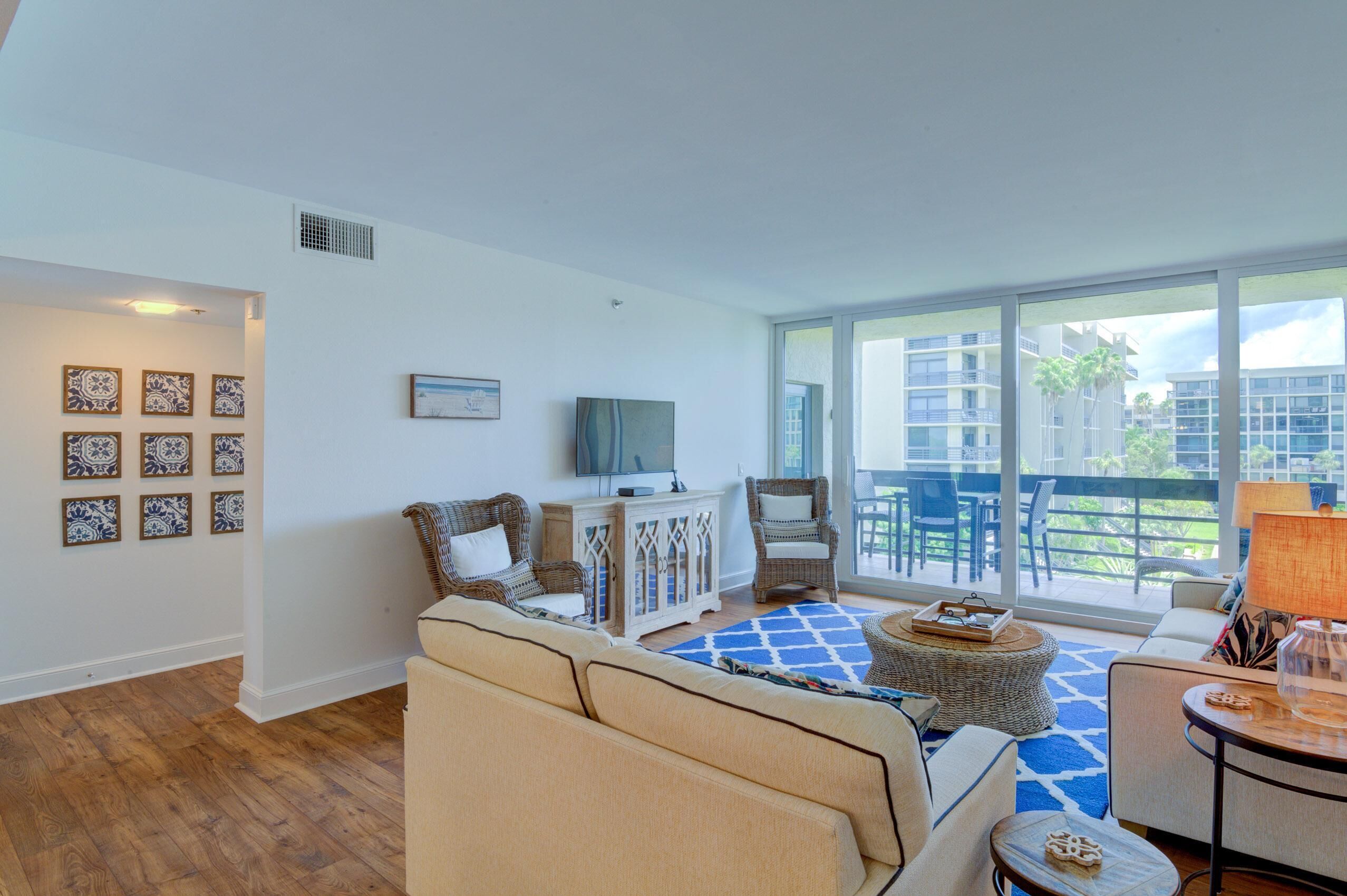 A spacious living room with a large window, a flat-screen TV, wicker furniture, and decorative tiles on the wall.