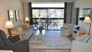 A modern living room with a sofa, armchair, and a small table with a plant. There is a balcony with chairs and a table, and a large window with curtains. - Partial Beach View 2 Bedroom Condo (Longboat Key)