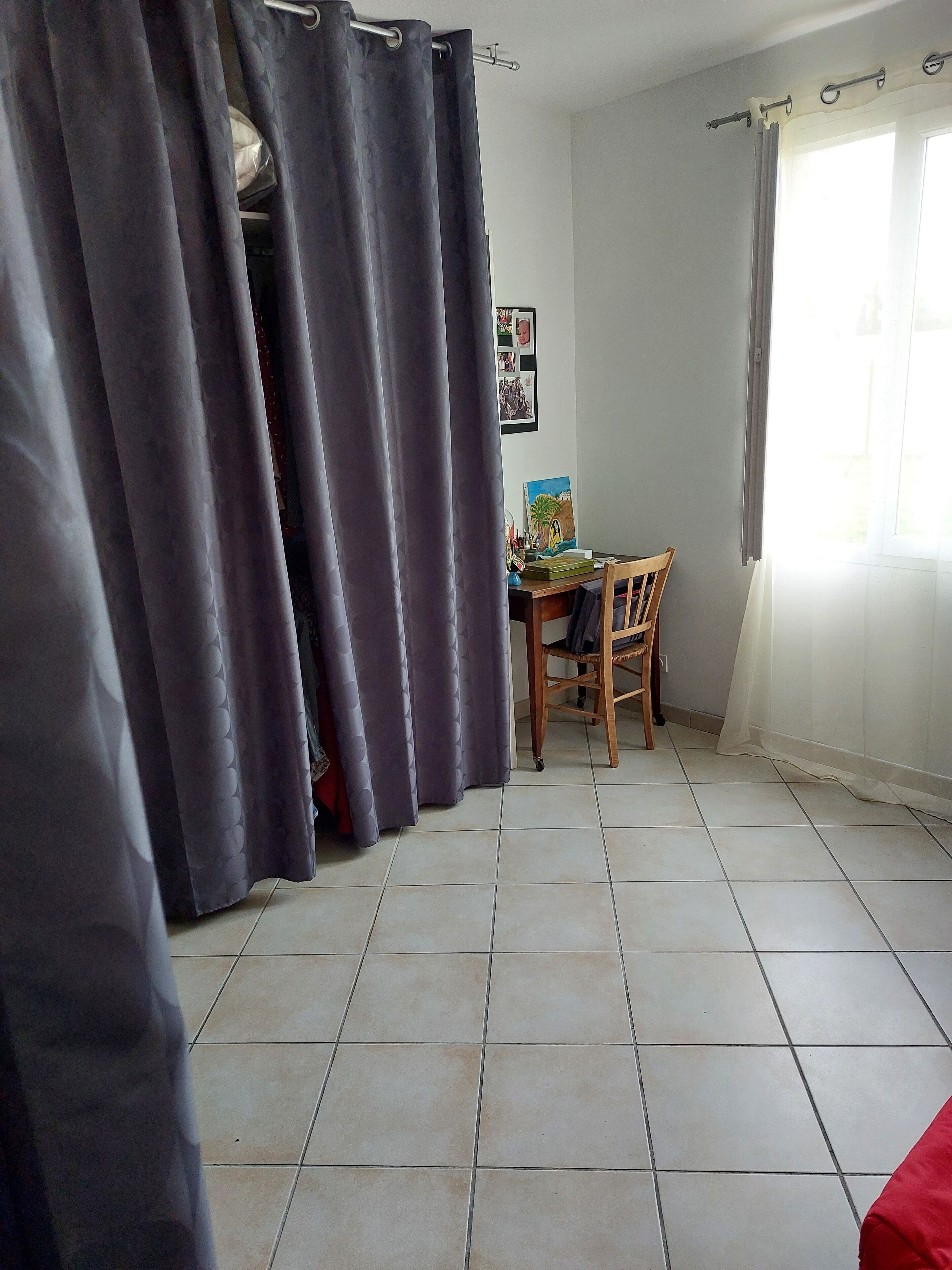 Une pièce avec un sol carrelé, une chaise en bois, une table sur laquelle est posée une peinture, et une fenêtre avec des rideaux.