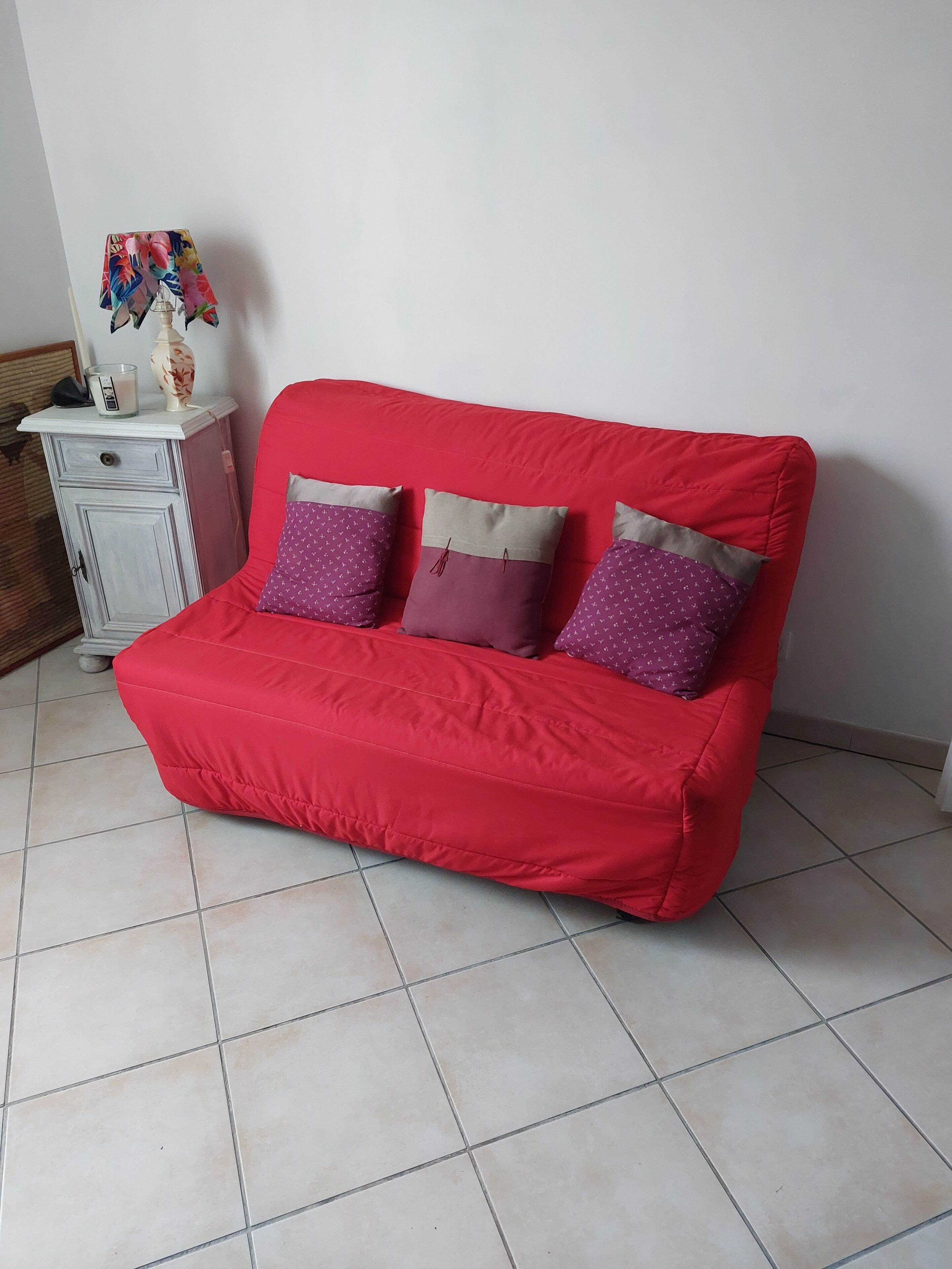 Ein rotes Sofa mit drei gemusterten Kissen, eine weiße Kommode mit einer Blumenvase und ein Fliesenboden.
