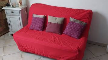 Ein rotes Sofa mit drei gemusterten Kissen, eine weiße Kommode mit einer Blumenvase und ein Fliesenboden.
