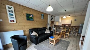 Una sala de estar con un sofá de cuero negro, una mesa de centro de madera y una alfombra gris. Hay dos sillones negros y una mesa de comedor de madera con sillas. La habitación tiene paredes de madera y un suelo de baldosas.