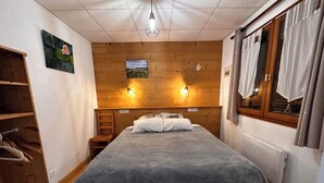 Un dormitorio con cabecera de madera, cama con manta gris, estante de madera con ropa y ventana con cortinas.