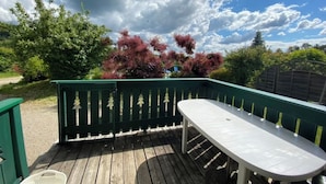 A wooden deck with a white table and chairs, surrounded by green fencing and trees. - Chalet des planchettes le lynx (Marigny)