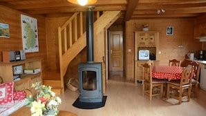 A wooden interior with a central wood-burning stove, a dining area with a red tablecloth, and a kitchen with wooden cabinets. - Chalet des planchettes le lynx (Marigny)