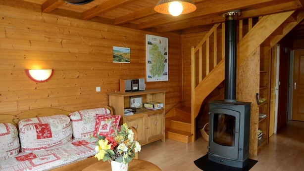 A cozy cabin interior with a wooden interior, a sofa with patterned cushions, a small table with a vase of flowers, a wooden cabinet, a map on the wall, and a wood-burning stove. - Chalet des planchettes le lynx (Marigny)