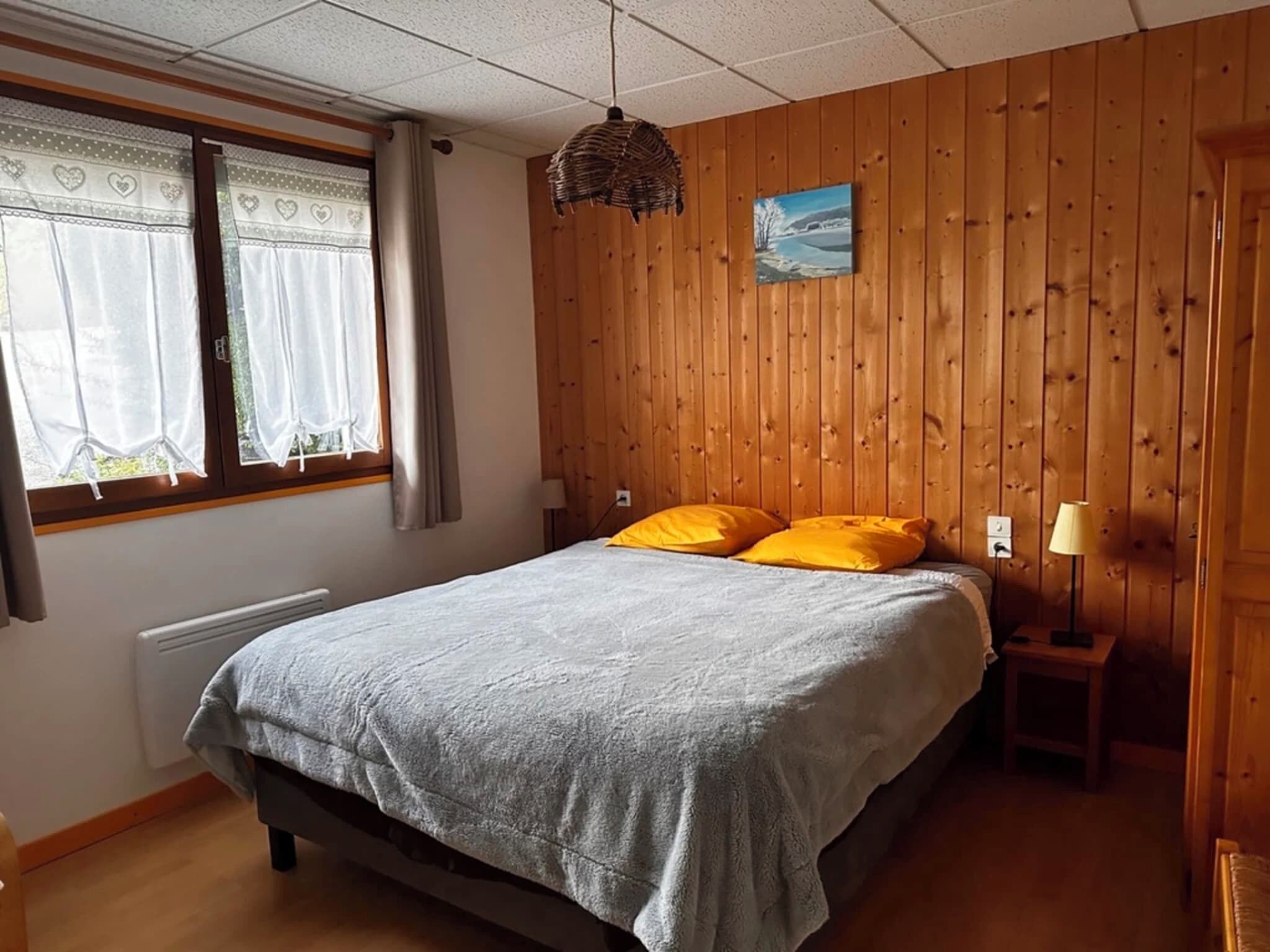 Ett sovrum med en säng, två kuddar, ett fönster med gardiner, en tavla som hänger på väggen och en sänglampa.