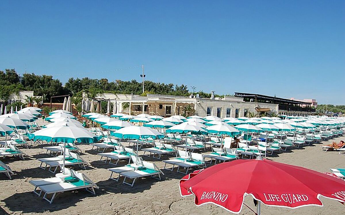 Una playa con filas de tumbonas y sombrillas, un edificio al fondo y una sombrilla roja con el texto 'LIFE GUARD'.