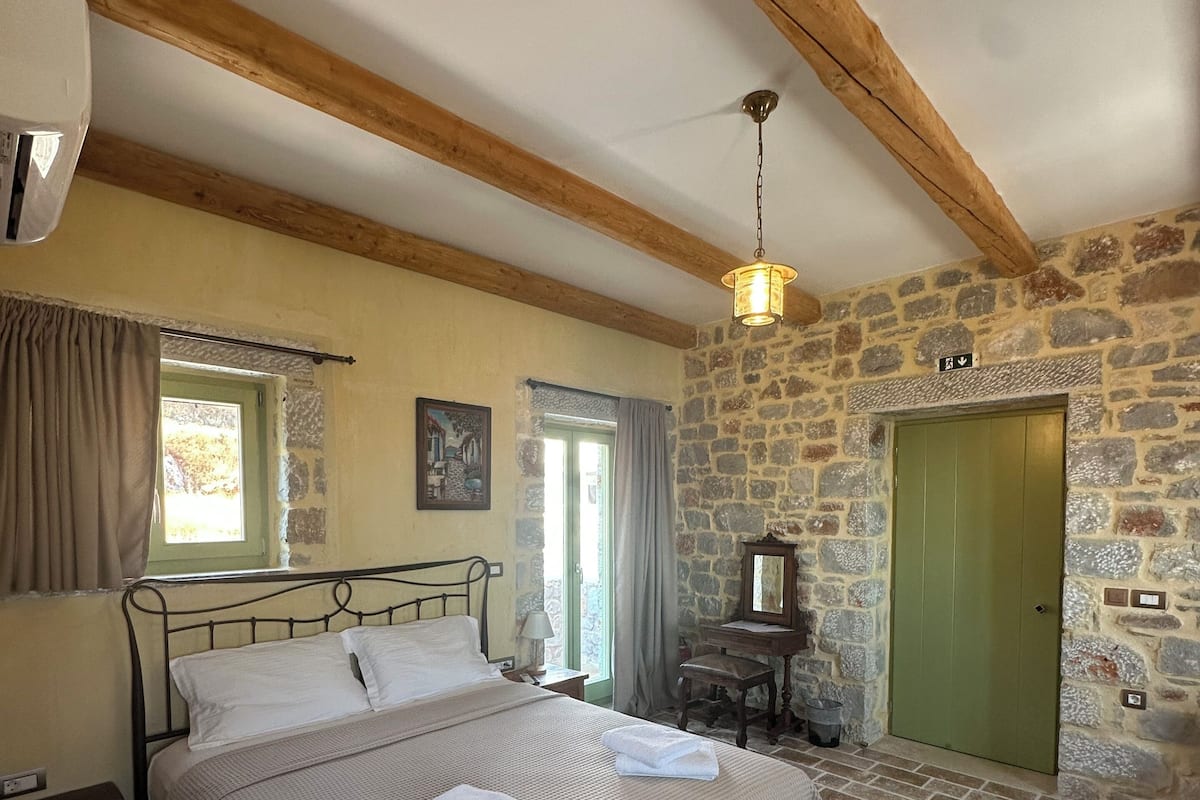 Ein Schlafzimmer mit einem Bett, einem Nachttisch, einer Kommode, einem Spiegel, einer Steinwand, einer Holzbalkendecke und einer grünen Tür.