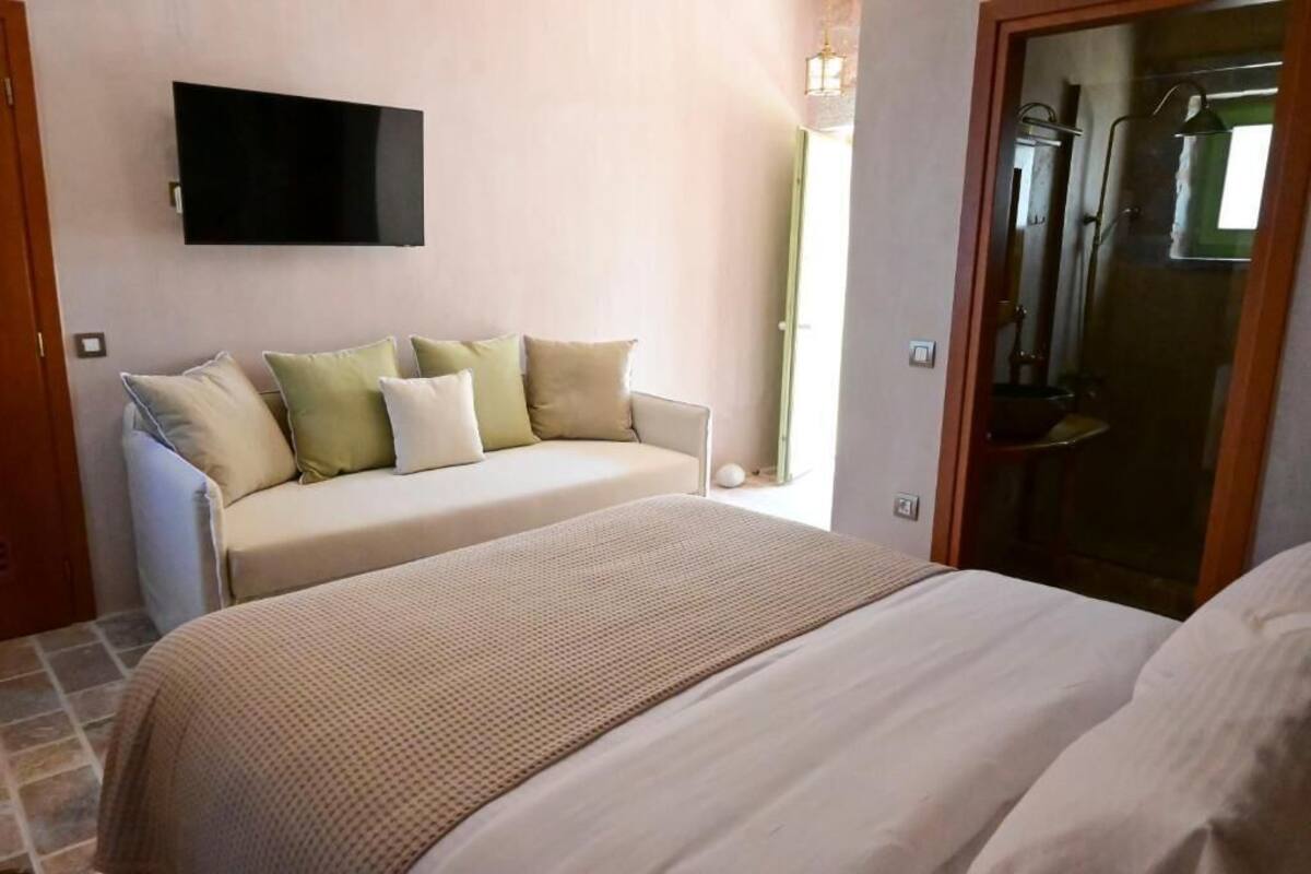 Ein Schlafzimmer mit einem Bett, einem Sofa, einem an der Wand montierten Fernseher und einem durch eine offene Tür sichtbaren Badezimmer.