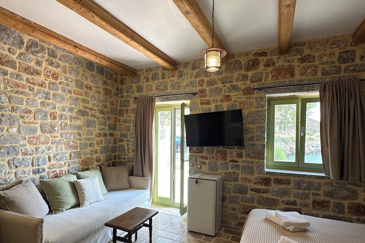 Ein gemütliches Zimmer mit Steinwänden, Holzbalkendecke, einer Couch, einem Bett, einem Fernseher und einem kleinen Kühlschrank.