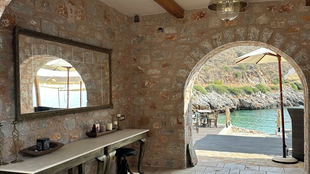 Habitació amb parets de pedra, sostre de fusta, una taula llarga amb tamborets i vista a una piscina a través d'una porta d'arc.