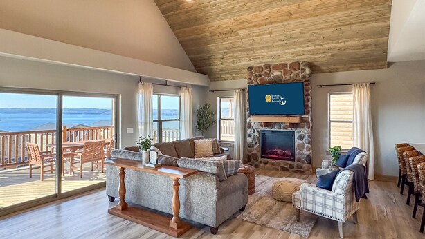 A spacious living room with a stone fireplace, a television mounted above it, and a balcony overlooking a body of water.