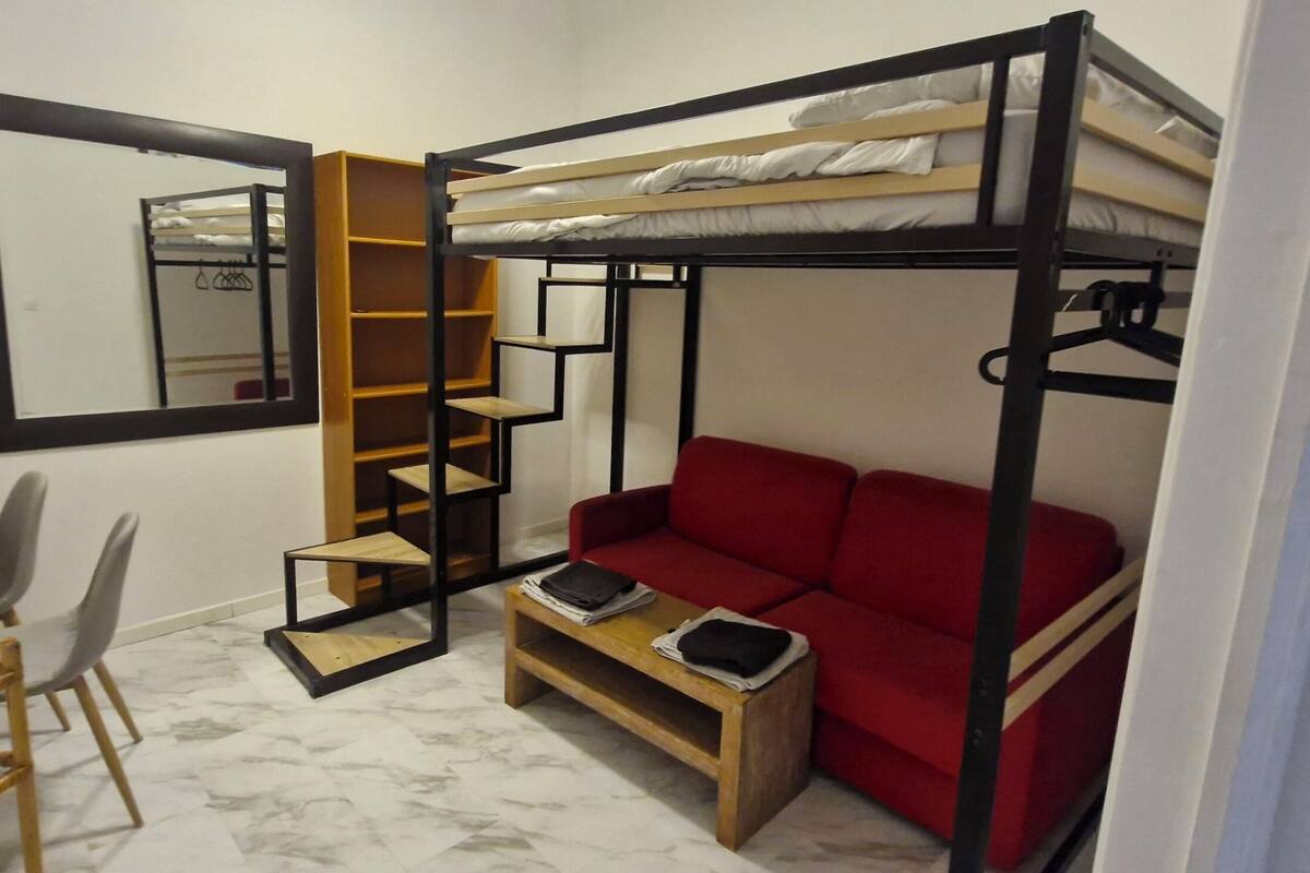 Quarto compacto com cama beliche, sofá vermelho, escrivaninha de madeira e uma cadeira.