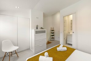 Ein modernes Hotelzimmer mit einem weißen Kleiderschrank, einem weißen Sessel, einem Bett mit senfgelber Bettdecke und einer kleinen Pflanze auf dem Nachttisch.