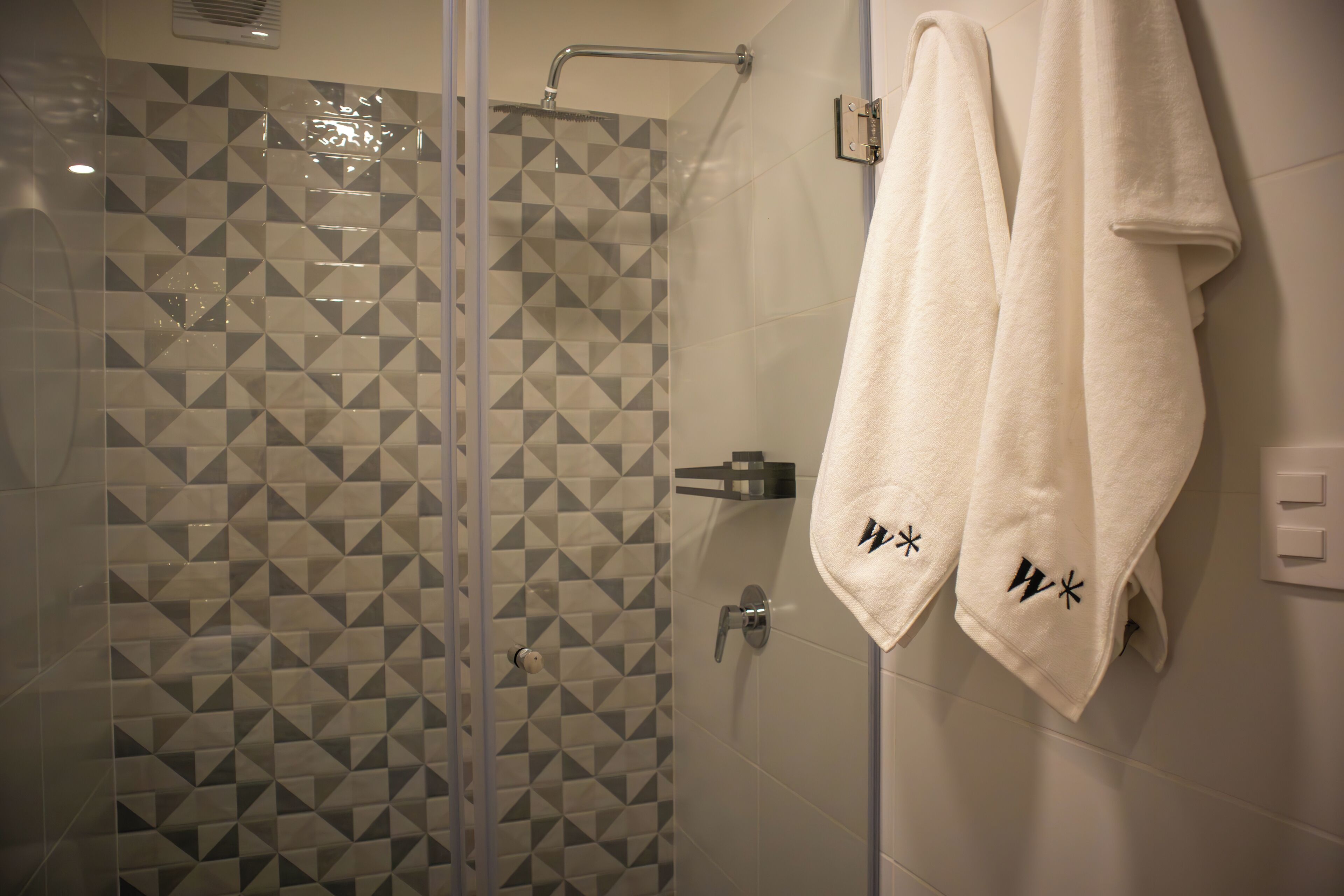 A shower with geometric tile pattern and two white towels with embroidered initials on a wall.