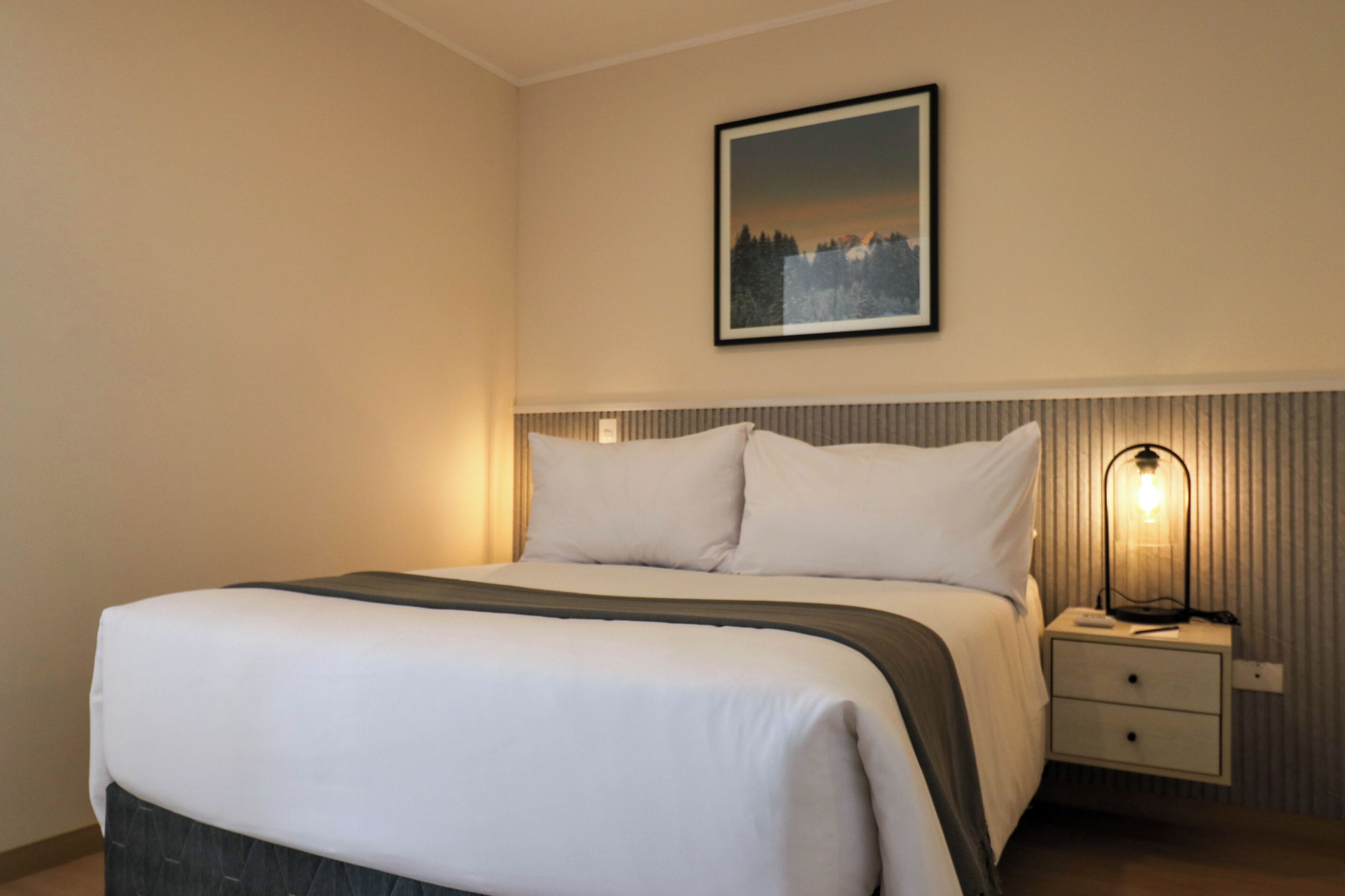 A neatly made bed with white linens and a dark throw blanket, a bedside lamp, and a framed picture on the wall.