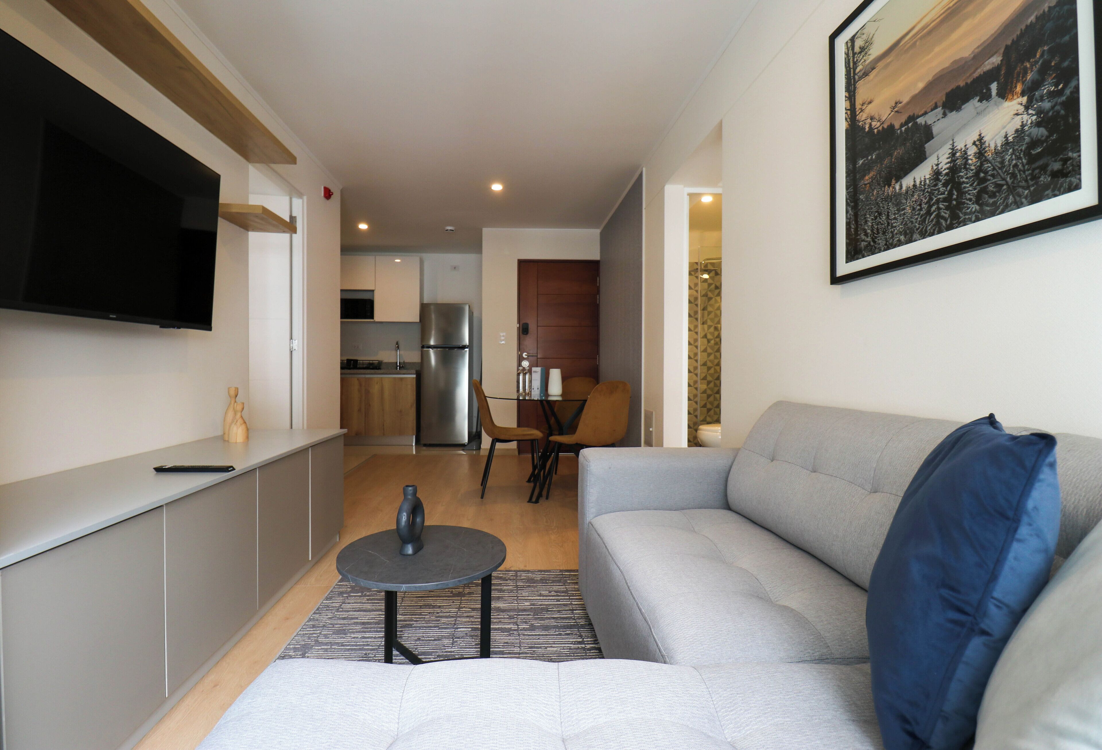 Ein modernes Wohnzimmer mit grauem Sofa, einem Wand-Fernseher, einem Essbereich mit Tisch und Stühlen sowie einer Küche im Hintergrund.