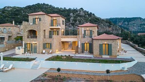 Una villa de piedra con varios niveles, mobiliario exterior y vistas a un paisaje montañoso.