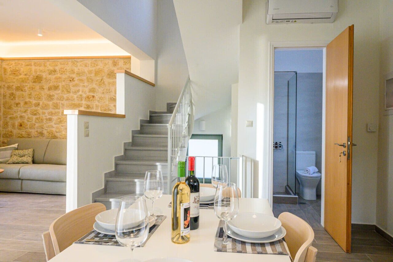 A dining area with a table set for four, a staircase, a stone wall, and a bathroom visible through an open door.