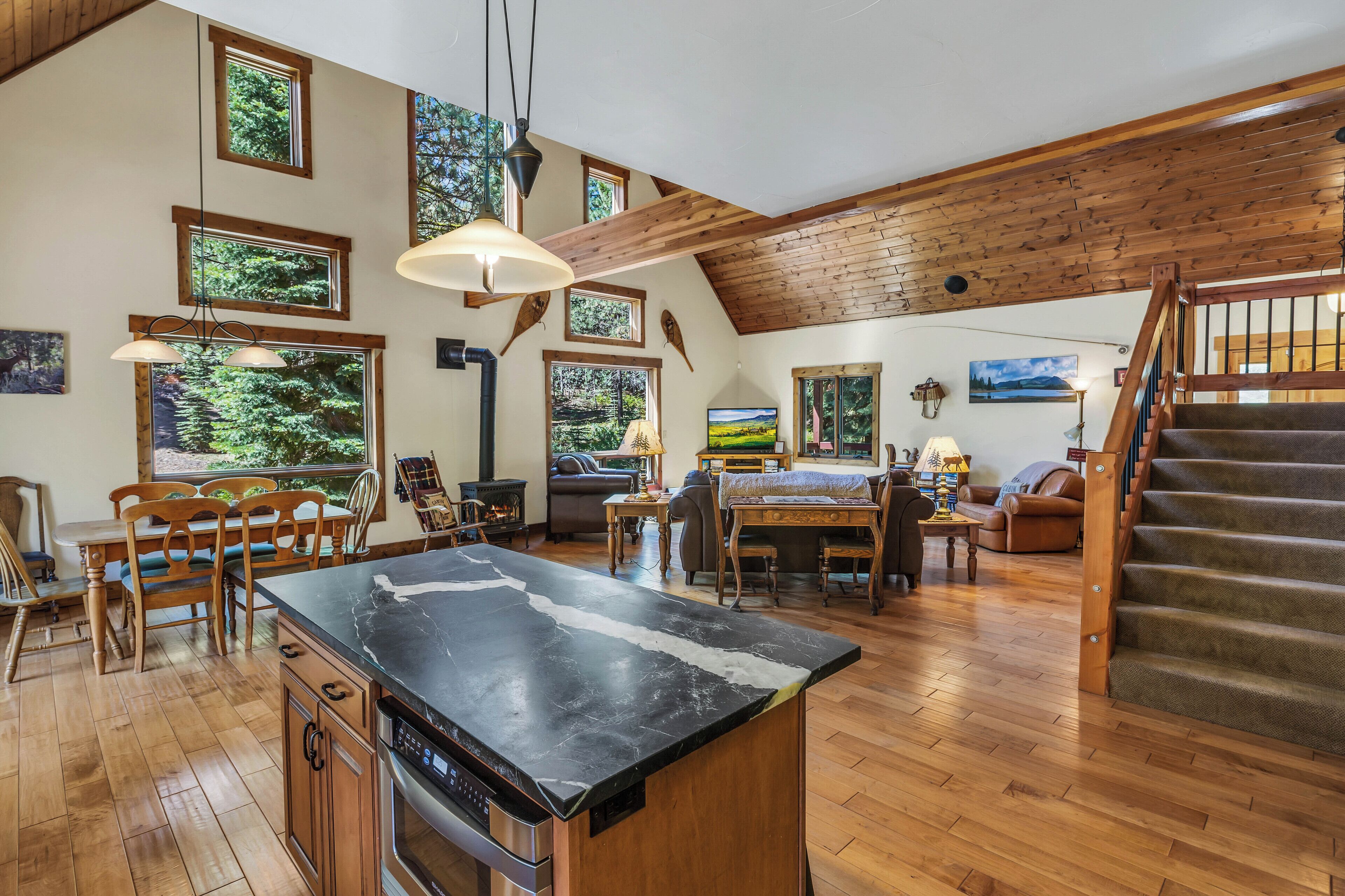 A spacious living area with a wooden staircase, large windows, and a central island with a black countertop.
