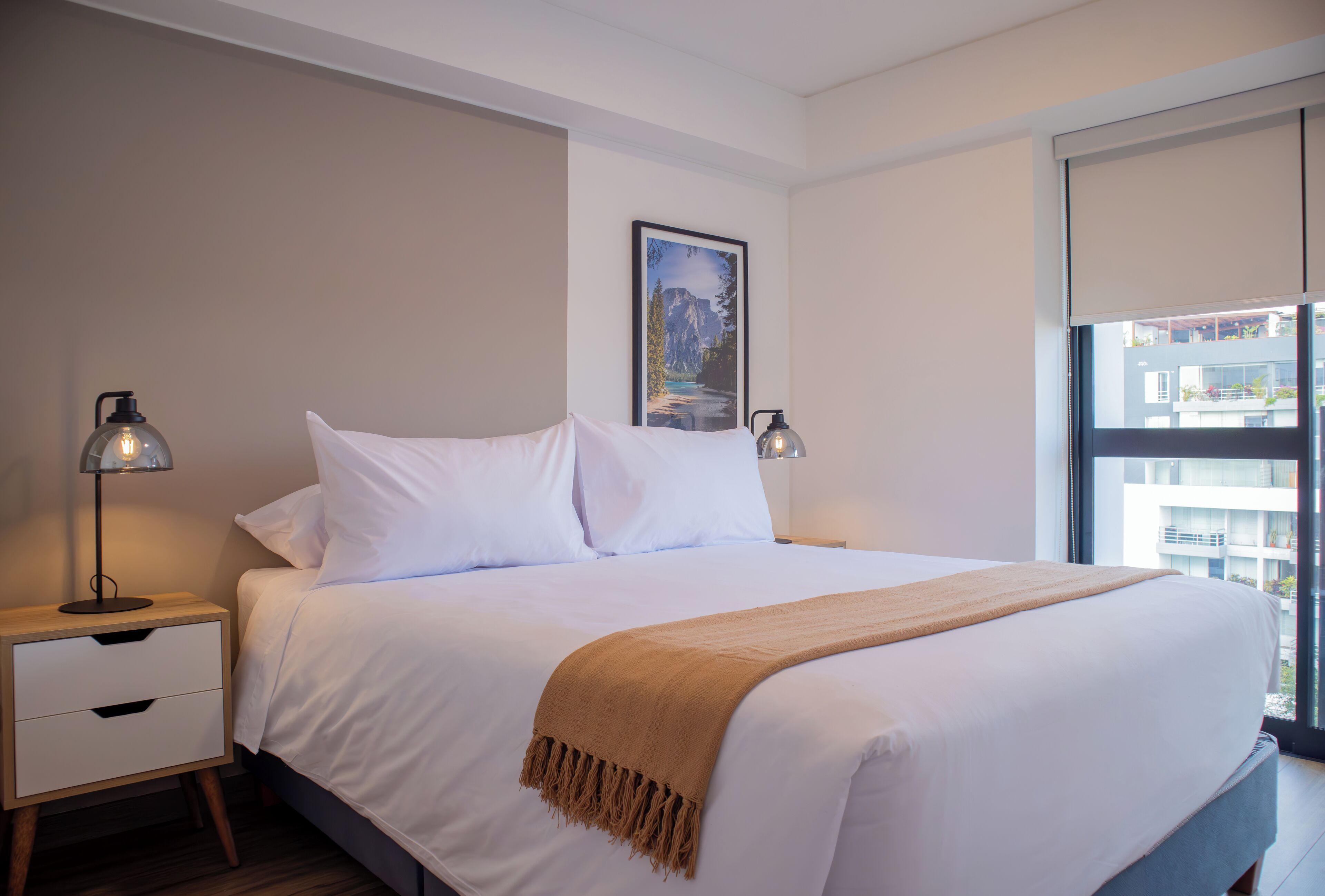 Una cama bien hecha con sábanas blancas y una manta marrón, dos lámparas de noche, un cuadro en la pared y una ventana con vistas a edificios.
